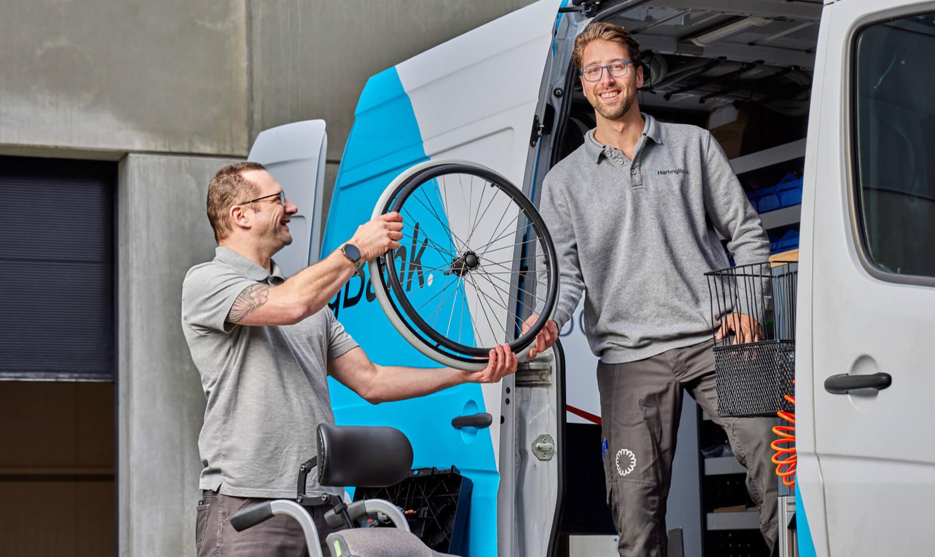 Een HartingBank bestelbus met 2 medewerkers en een rolstoel. Een persoon handigt een rolstoelwiel over aan de ander.